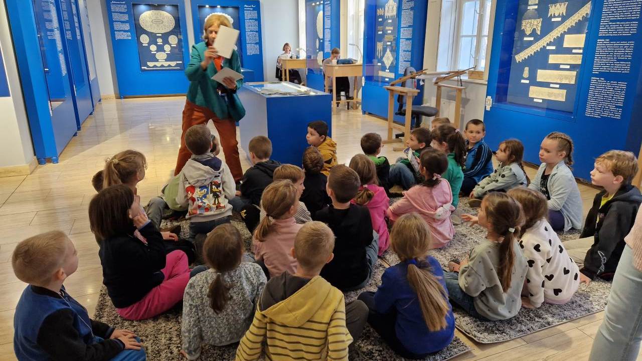 Gyerekek csoportja múzeumpedagógiai foglalkozáson ül és figyel.